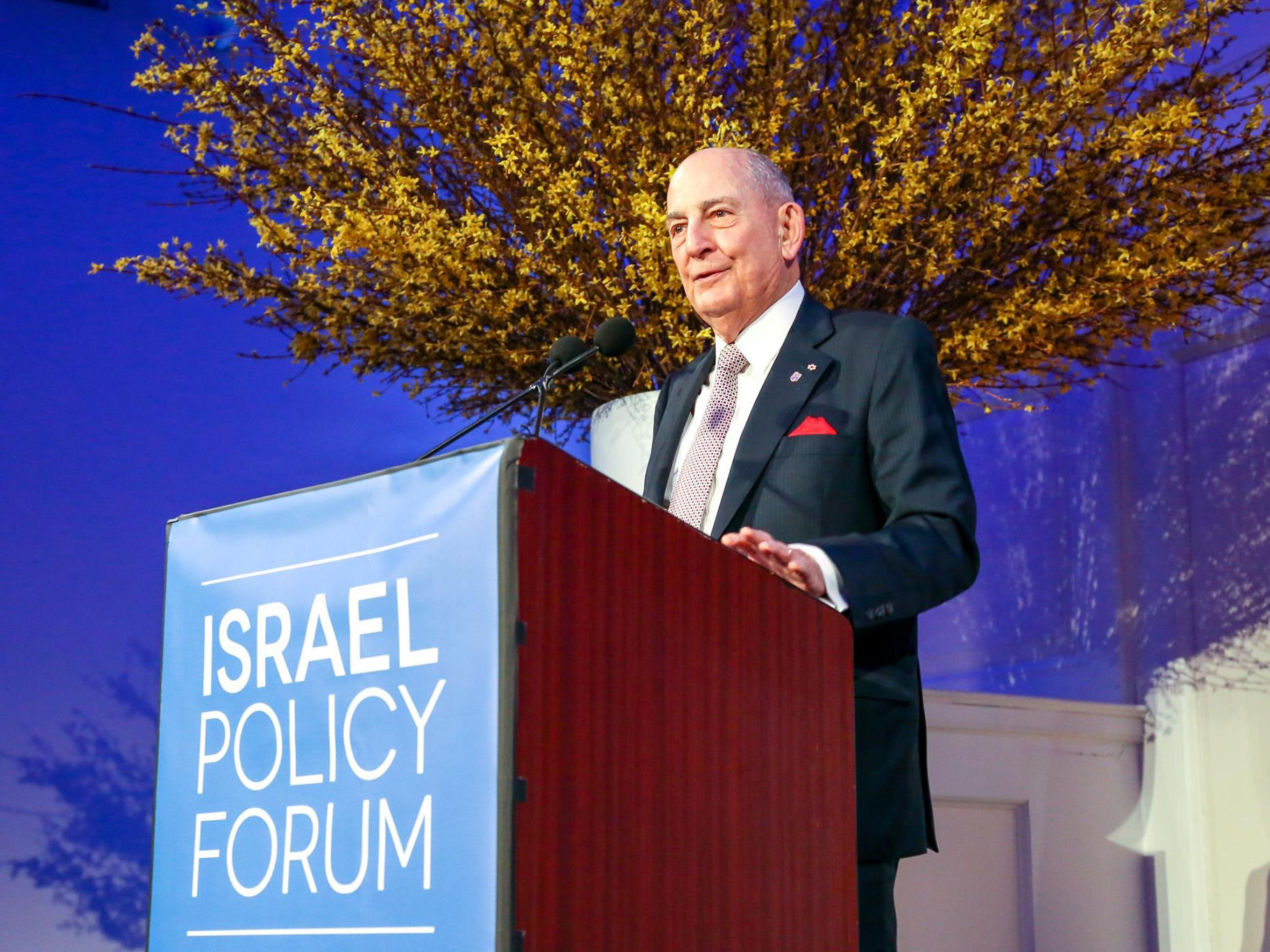 An older man in a suit speaks at a podium with a sign that reads Israel Policy Forum. Yellow branches are arranged behind him, and the background is illuminated with blue lighting.