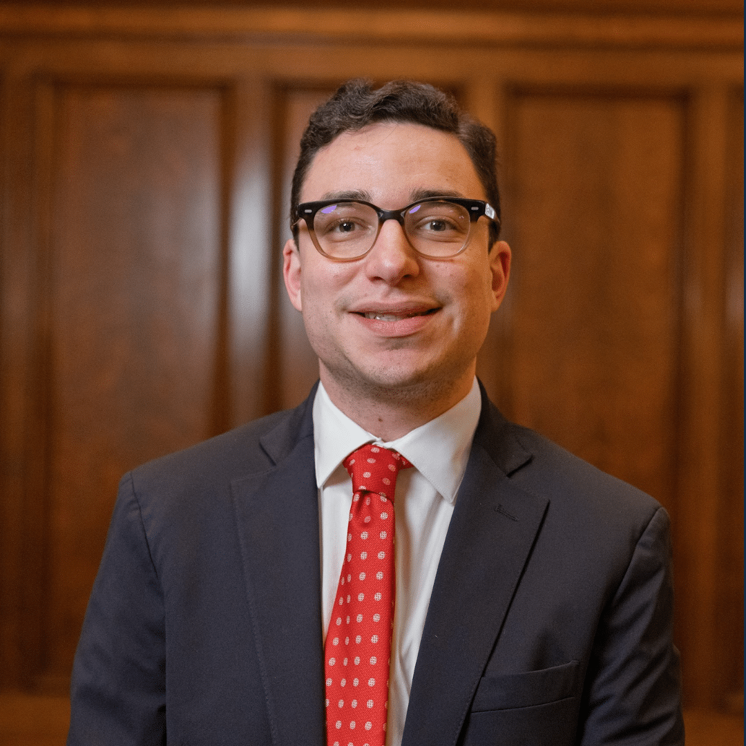 A man wearing glasses, a dark suit, white shirt, and red polka dot tie stands in front of a wooden paneled wall, smiling at the camera.