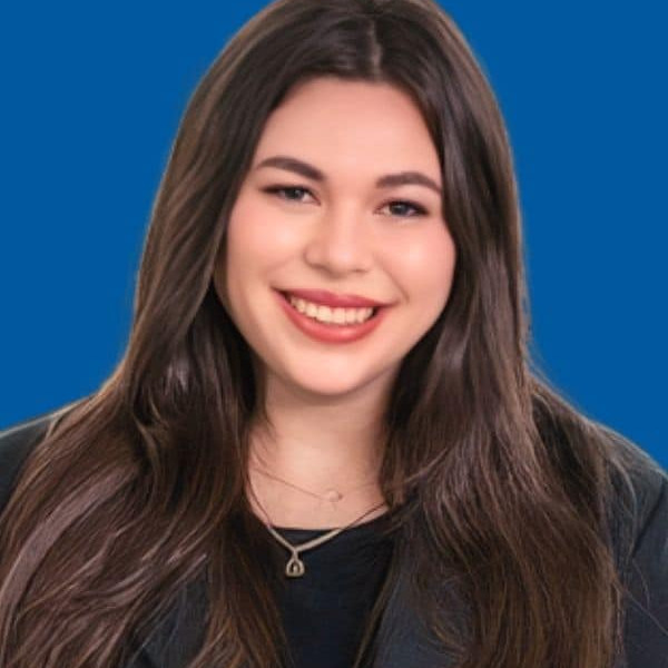 A young woman with long brown hair, wearing a black top and blazer, smiles against a solid blue background.