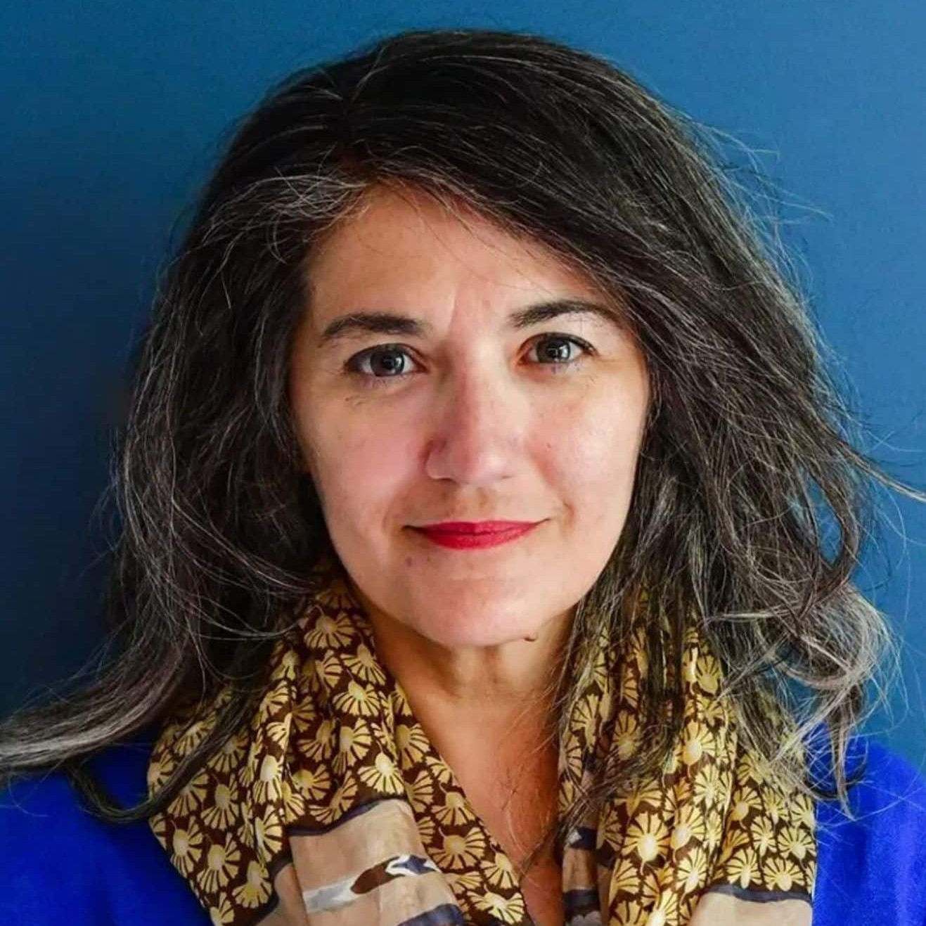 A woman with long, wavy brown and gray hair stands in front of a blue background. She is wearing a blue top and a patterned scarf with gold, brown, and blue colors. She is looking at the camera and smiling slightly.