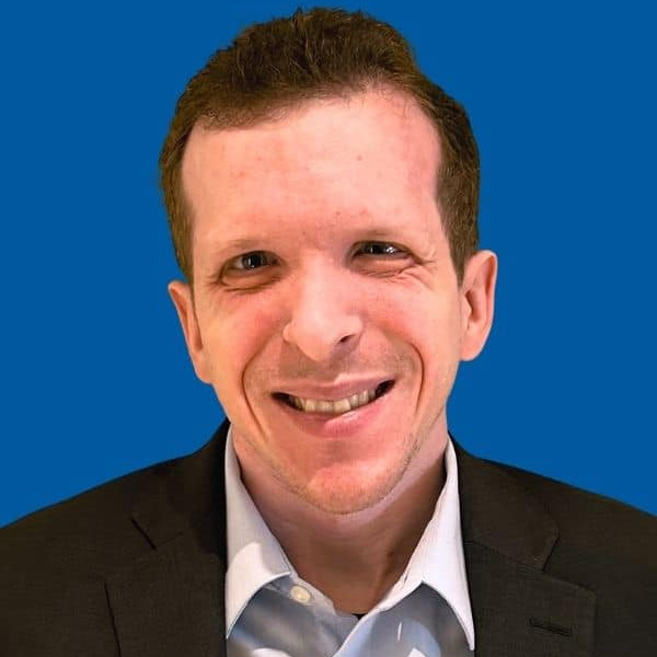 A man with short hair, wearing a dark suit jacket and a light blue collared shirt, smiles at the camera against a solid blue background.
