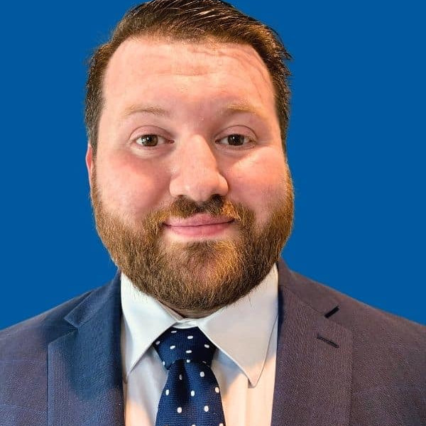 A man with short brown hair and a beard wearing a blue suit, white shirt, and blue tie with white polka dots, posing in front of a solid blue background.