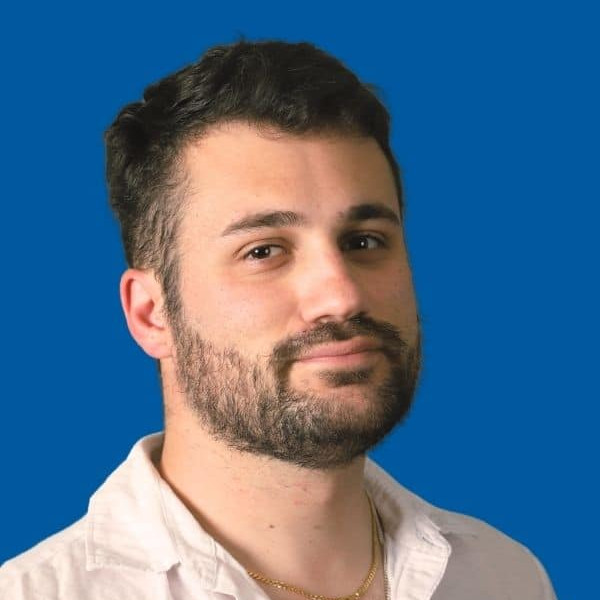 A man with short dark curly hair and a beard, wearing a white shirt and a thin gold necklace, poses against a solid blue background.