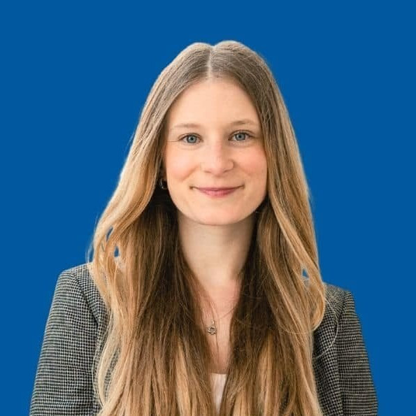 Woman with long blonde hair, wearing a checkered blazer and white shirt, smiling in front of a solid blue background.
