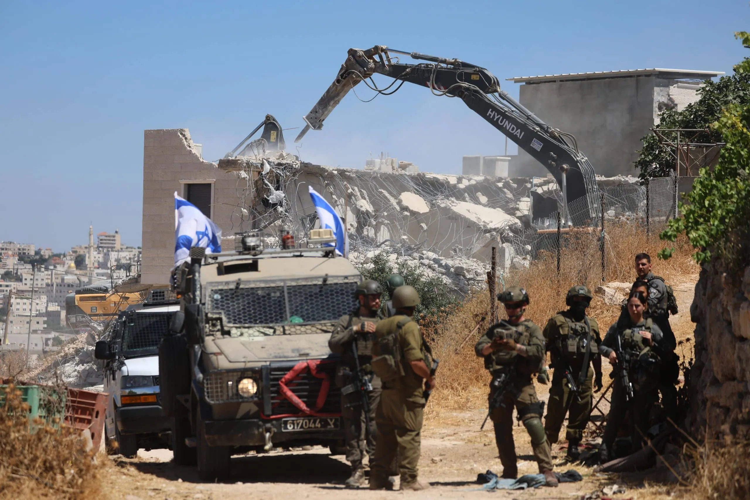 Israeli soldiers stand near armored vehicles with flags, as a construction machine demolishes a building in a dry, urban area under a clear sky. Dust and debris surround the partially destroyed structure.