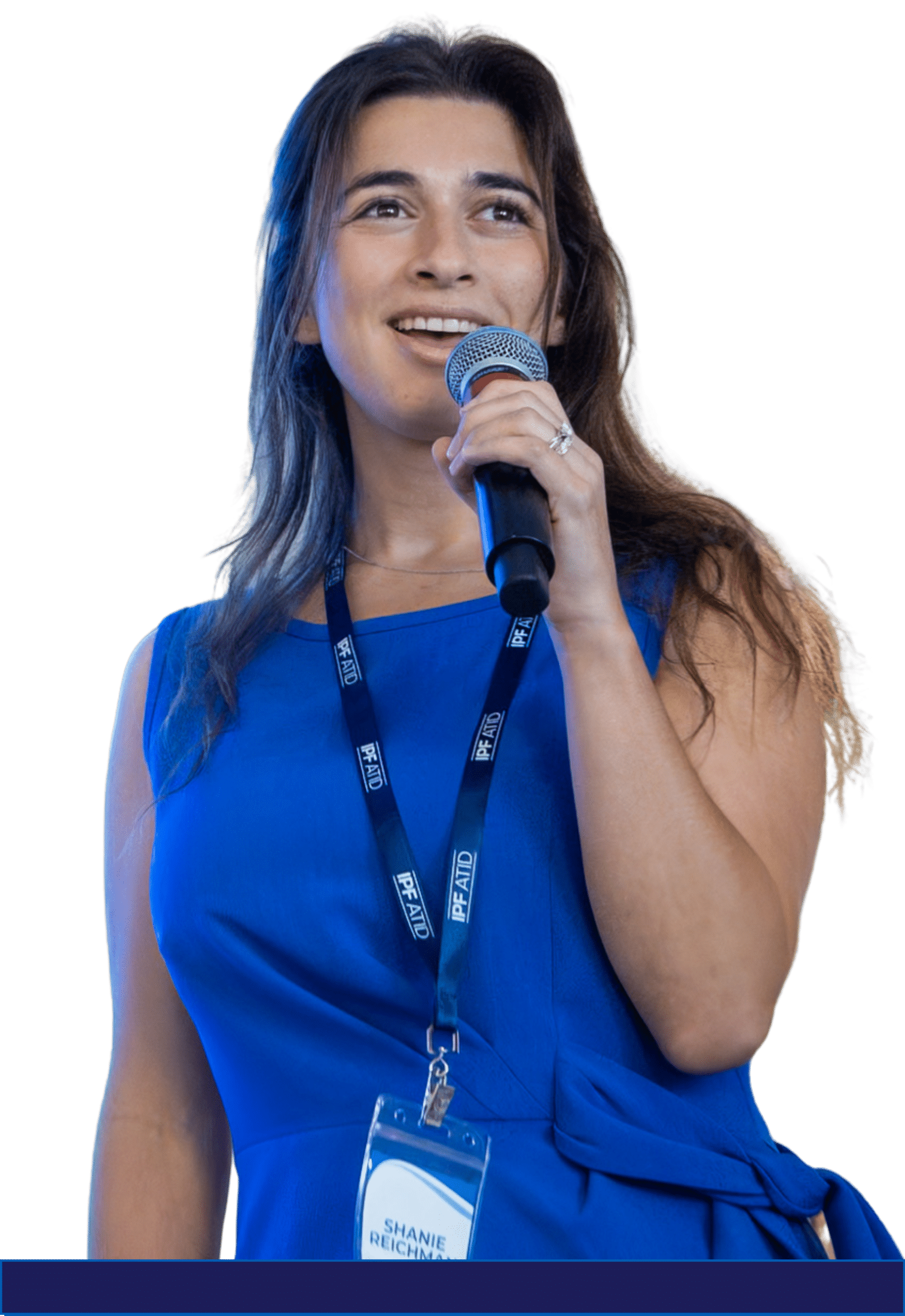 A woman in a blue dress speaks into a microphone, wearing a conference lanyard and name badge, smiling and looking slightly to the side.
