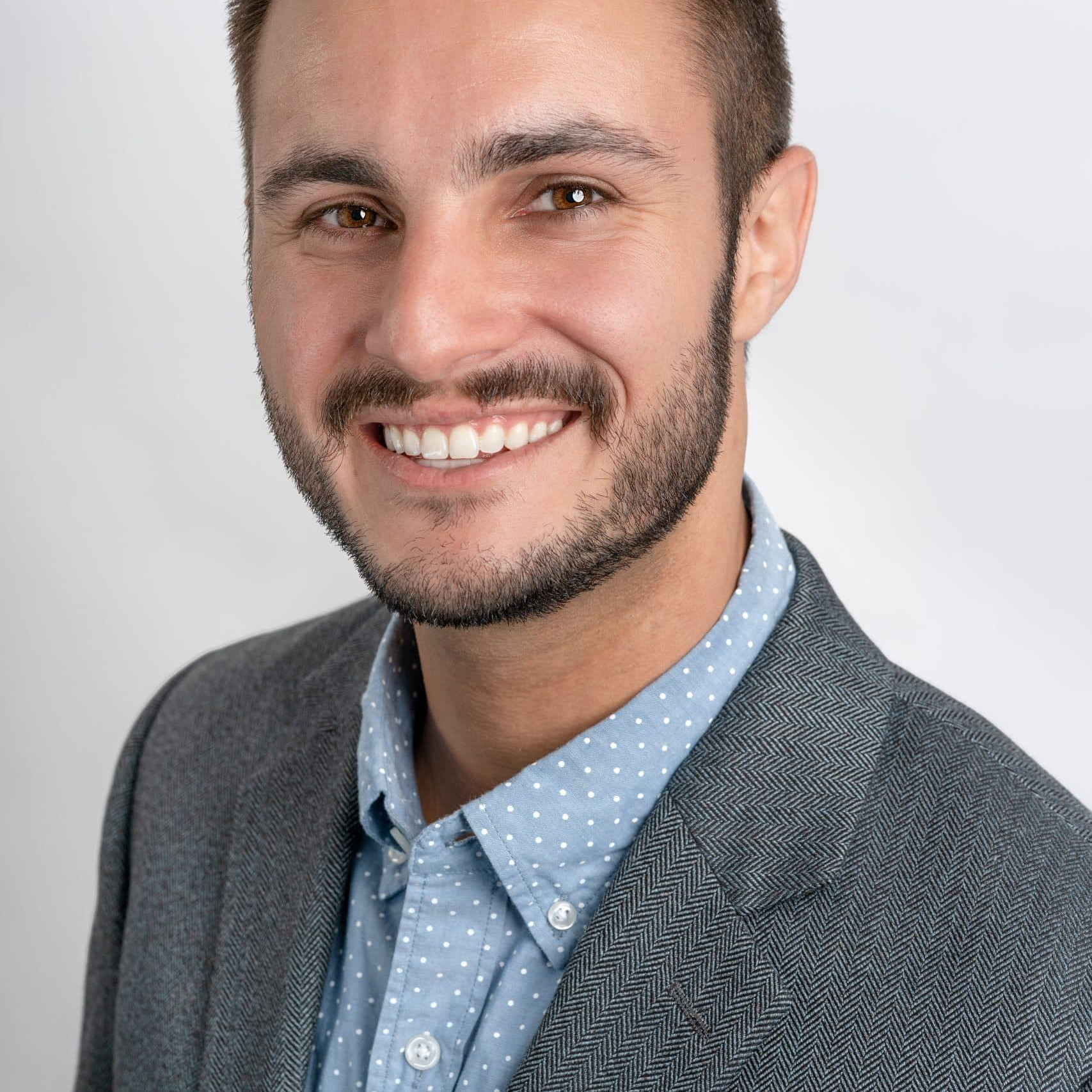 A smiling man with a short beard wears an atid grey blazer over a blue polka-dot shirt.