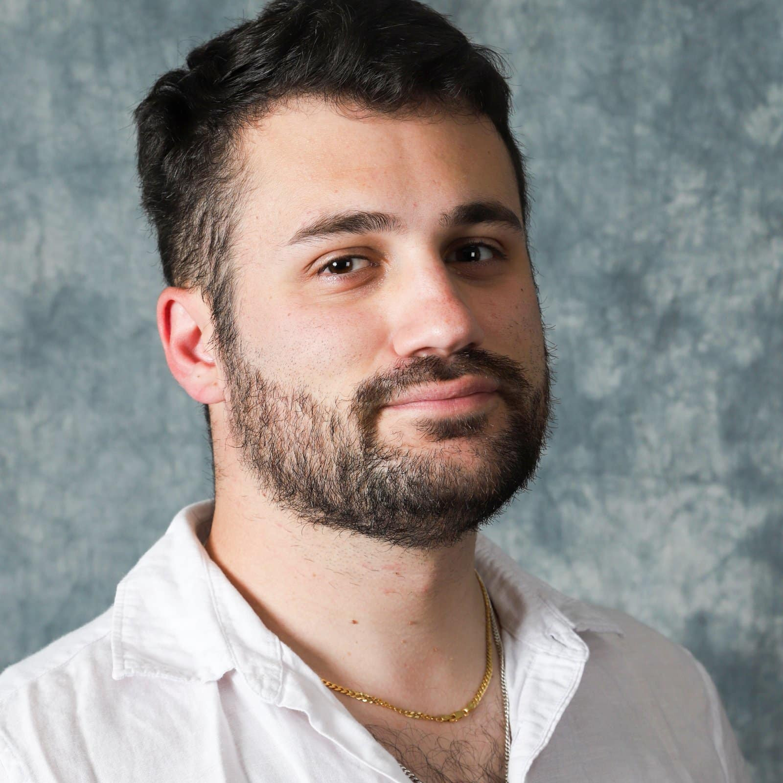 Headshot of an Atid man with short dark hair and a beard, wearing a white collared shirt and a gold chain.